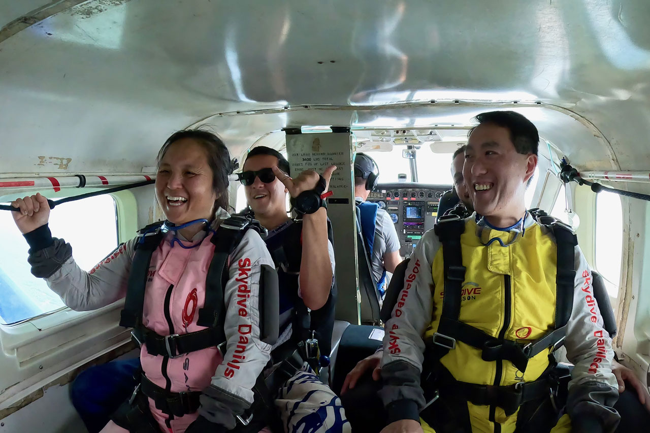Three people in skydiving suits smile and hold onto bars inside a small airplane, preparing for a jump. The cockpit and pilot are visible in the background. The atmosphere appears lighthearted and excited.