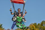 Two men tandem skydiving, descending with a red parachute against a clear blue sky. The front man wears a green jumpsuit and goggles, while the instructor behind him wears sunglasses. Trees with autumn leaves are visible below.