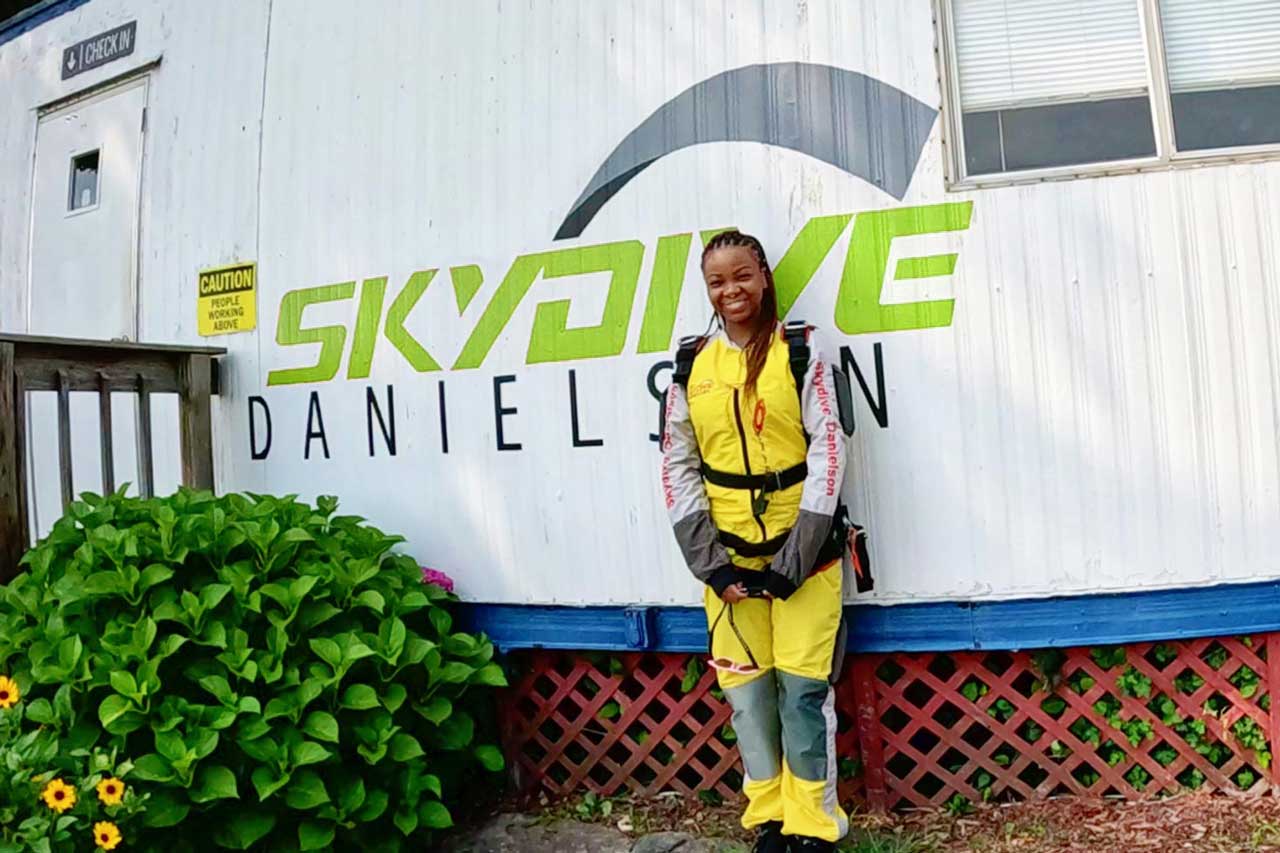 A person wearing a yellow jumpsuit and harness stands smiling in front of a white building with the words "Skydive Danielson" painted on it. Green plants and yellow flowers grow nearby.