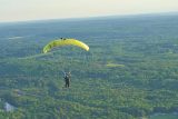 A person is parachuting with a bright yellow parachute above a vast, green, forested landscape, with fields, trees, and a river visible below under a clear sky.