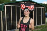 A smiling young woman stands outside a building with a “Cookie” sign, giving two thumbs up. She has a blue star painted on her forehead and is wearing a tank top that says “STAY THE” in pink letters.