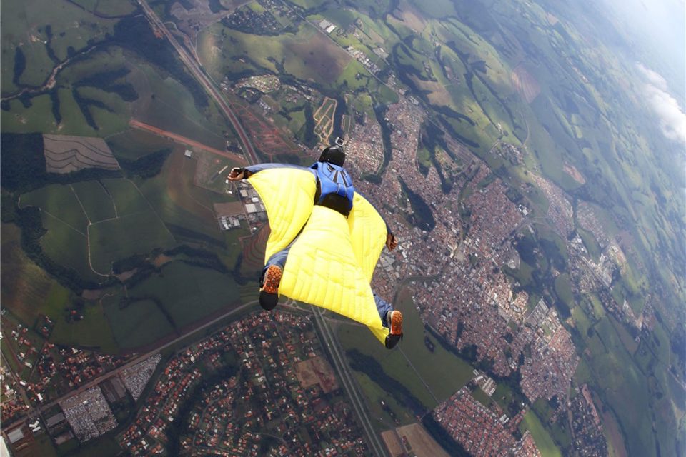 A person in a yellow and blue wingsuit is flying high above a landscape of green fields, roads, and towns, captured from behind during a wingsuit flight.